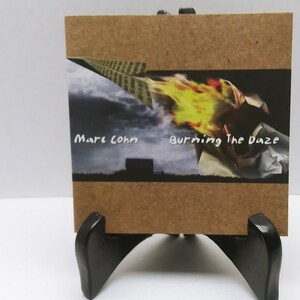 May include: A brown cardboard CD case with a black and white image of a building and a yellow flame. The text "Marc Cohn" and "Burning The Daze" is printed on the case.