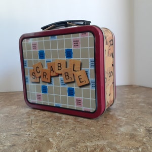 May include: A red metal lunchbox with a Scrabble game board design. The lid features the word "SCRABBLE" spelled out with letter tiles.