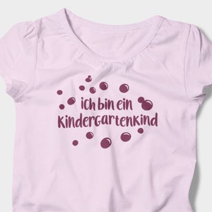 Könnte beinhalten: Ein hellrosa T-Shirt mit kurzen Ärmeln und rundem Ausschnitt. Auf dem Shirt steht der Text "ich bin ein Kindergartenkind" in Dunkelviolett, umgeben von violetten Punkten. Eine Jeans-Shorts und ein weißer Sneaker sind zu sehen.