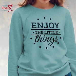 Könnte beinhalten: Ein hellblauer Pullover mit dem Aufdruck "ENJOY THE LITTLE things" in Schwarz auf der Vorderseite. Der Text ist von einem Kreis aus kleinen schwarzen Punkten umgeben.