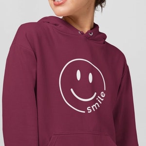 Könnte beinhalten: Burgunderfarbener Hoodie mit einem weißen Smiley und dem Wort "smile" darunter. Der Hoodie hat eine Kapuze mit Kordelzug und eine Kängurutasche.