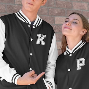 Könnte beinhalten: Zwei schwarze und weiße Baseballjacken mit weißen Buchstaben. Die Jacke links hat ein weißes "K" auf der Brust. Die Jacke rechts hat ein weißes "P" auf der Brust.