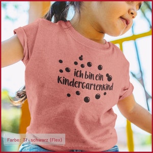 Könnte beinhalten: Ein pinkfarbenes T-Shirt mit schwarzem Text, der "ich bin ein kindergartenkind" lautet, umgeben von schwarzen Kreisen. Der Text ist in Deutsch und bedeutet "Ich bin ein Kindergartenkind".
