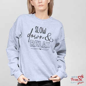 Könnte beinhalten: Grauer Pullover mit dem Text "SLOW down & RELAX" in schwarzer Schrift. Der Text ist in handschriftlicher Form. Der Pullover hat einen Rundhalsausschnitt und lange Ärmel.