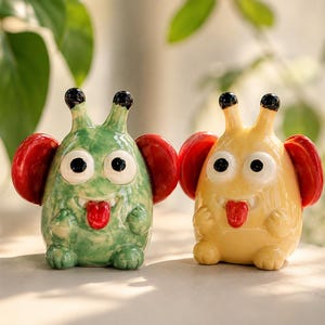 May include: Two ceramic figurines shaped like small creatures, one green and one yellow. Both have large white eyes with black pupils, two antennae with black tips, red ears, and a red tongue sticking out from a small mouth with visible teeth.