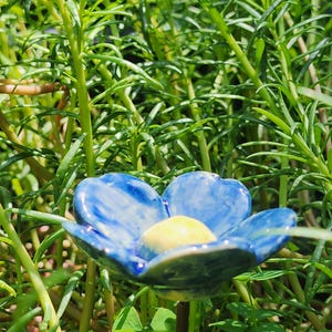 Peut inclure: Une fleur en céramique composée de cinq pétales arrondis avec un vernis marbré bleu et blanc brillant et un centre jaune lisse et surélevé. La fleur est fixée sur une fine tige brun foncé, positionnée au milieu d'un feuillage vert vif.