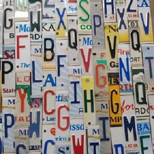 May include: A close-up of a wall art piece made from a collection of vintage license plates. The plates are arranged vertically and feature a variety of letters, numbers, and words. The letters are painted in different colors, including red, blue, green, and yellow. The letters are arranged in a random order, creating a unique and abstract design.