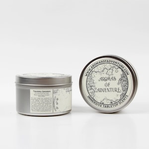 May include: Two silver-colored metal tins. One tin has a label that reads "Training Grounds" and the other has a lid with the text "Aromas of Adventure" and a map design. The tins are for immersive tabletop scents.