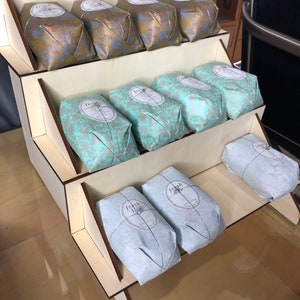 May include: A wooden display stand with three tiers, each holding wrapped soaps. The soaps are wrapped in gold and blue patterned paper with a circular label that says "fresh" on them.