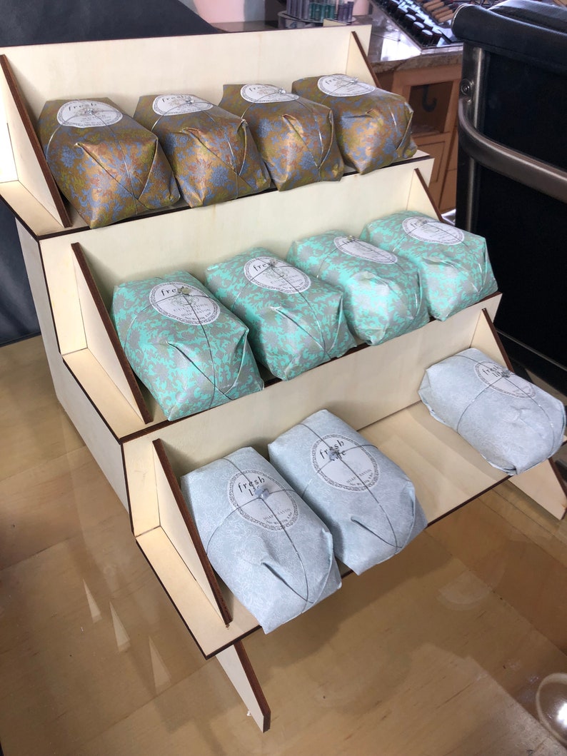 May include: A wooden display stand with three tiers, each holding wrapped soaps. The soaps are wrapped in gold and blue patterned paper with a circular label that says "fresh" on them.