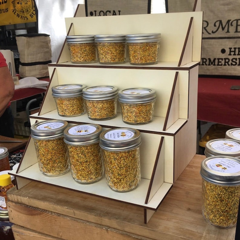 May include: A wooden display stand with three shelves holds multiple glass jars filled with a yellow substance. The jars have silver lids and white labels. The stand is labelled "Local" and "Farmers Market".