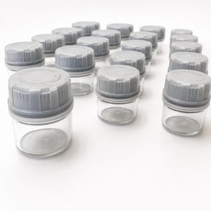 May include: A collection of small, clear glass jars with grey plastic screw-on lids. Each lid features a textured edge and embossed text that reads "PUSH DOWN & TURN" and "CLOSE TIGHT". The jars are arranged in a grid pattern on a white surface.