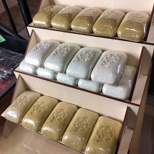 May include: A wooden display stand with three shelves holding multiple bars of green and white soap. The soap bars are wrapped in clear plastic and have a decorative pattern.