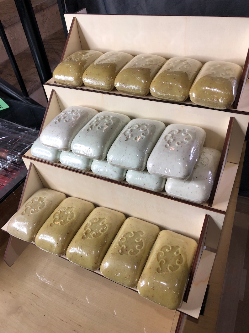 May include: A wooden display stand with three shelves holding multiple bars of green and white soap. The soap bars are wrapped in clear plastic and have a decorative pattern.