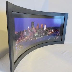 May include: A black curved frame with a nighttime cityscape photograph. The city skyline is lit up with lights and the river is dark.