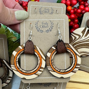 May include: A pair of wooden earrings with a brown leather accent. The earrings are round and have a swirl pattern in shades of brown and orange. The earrings are displayed on a brown card with the text "Truly Olive" printed on it.