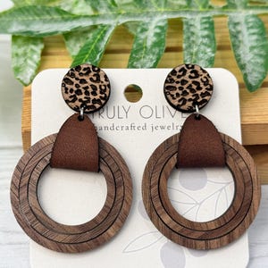 May include: A pair of brown wood and leather earrings with a leopard print design. The earrings are made of three layers of wood and have a brown leather accent. The earrings are labeled "TRULY OLIVE handcrafted jewelry".