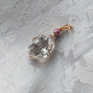 Könnte beinhalten: Goldener Anhänger mit einem großen, klaren, oval geschliffenen Edelstein. Ein kleiner, runder, rosa Edelstein ist über dem größeren Stein gesetzt. Der Anhänger wird auf einer weißen, strukturierten Oberfläche präsentiert.