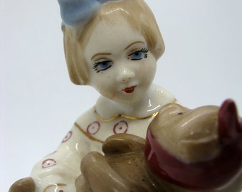 May include: A porcelain figurine of a young girl with blonde hair and a light blue bow. She is holding a brown object with a red band. The girl is wearing a white dress with red circles and gold trim.
