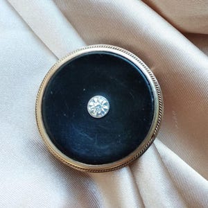 Könnte beinhalten: Vintage runde Brosche mit schwarzem Mittelteil und einem funkelnden, klaren Edelstein in der Mitte. Die Brosche hat einen goldfarbenen Metallrahmen mit einer seilartigen Umrandung. Der Hintergrund ist ein hellbrauner Stoff.