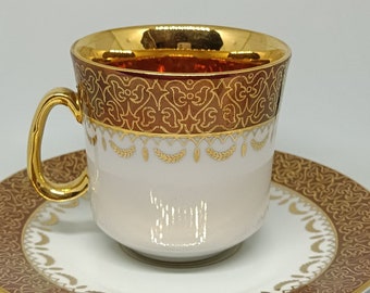 May include: A white porcelain teacup and saucer set with a gold rim and intricate brown and gold floral pattern.