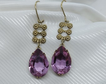 May include: A pair of vintage-style earrings featuring large, teardrop-shaped, light purple crystals. The earrings have a gold-colored metal design with floral accents and a hook closure. The earrings are displayed against a white, textured background.