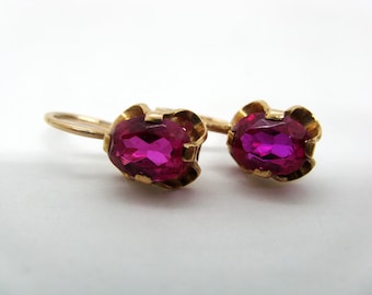 May include: A pair of gold-colored earrings featuring oval, magenta-colored gemstones. The gemstones are set in a decorative, scalloped gold setting. The earrings have a classic hook design.