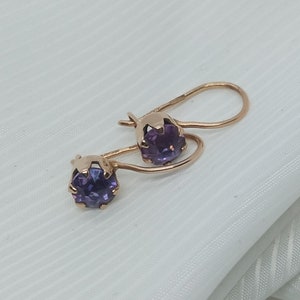 May include: A pair of gold-colored earrings with a lever back closure. Each earring features a round, deep purple gemstone. The earrings are displayed on a white, textured surface.