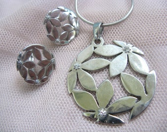 May include: A silver floral jewelry set featuring a pendant necklace, a pair of earrings, and a ring. The pendant and earrings have a flower design with small, clear gemstones. The ring has a similar floral design.
