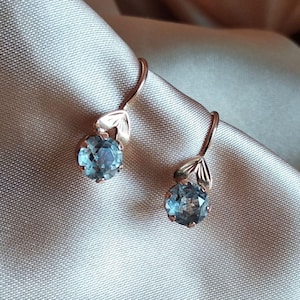 Könnte beinhalten: Ein Paar Ohrhänger aus Roségold mit runden, facettierten hellblauen Edelsteinen. Jeder Ohrring hängt an einem geschwungenen Drahtbügel und verfügt über ein kleines, poliertes, blattförmiges Metallelement direkt über der runden Steinfassung.