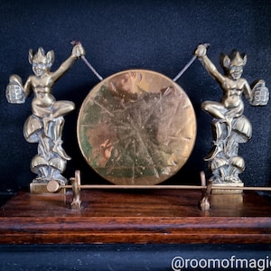 May include: A brass gong with two small brass figures holding a mallet on each side. The gong is mounted on a wooden base.