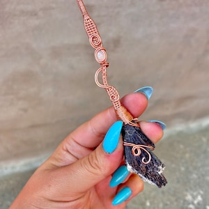 Puede incluir: Una escoba de bruja envuelta en alambre de cobre con un cristal blanco y negro y una piedra preciosa de ópalo rosa.