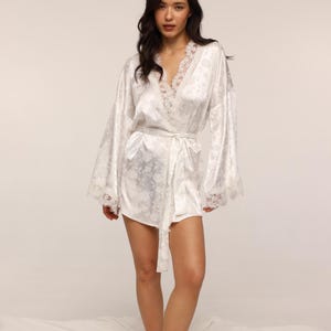 May include: A white satin robe with a floral pattern and lace trim. The robe has long, wide sleeves and a matching belt. The model is wearing the robe. The robe is a piece of sleepwear.