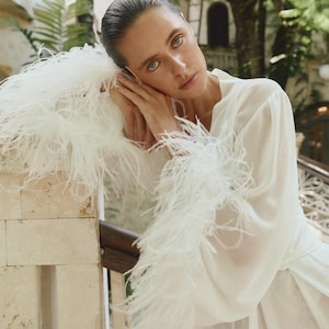 Pode incluir: Um roupão branco e fluido com acabamento de penas nas mangas. A pessoa está encostada a uma parede de pedra. O fundo inclui vegetação e uma escada, sugerindo um ambiente ao ar livre. O design do roupão é elegante e arejado.