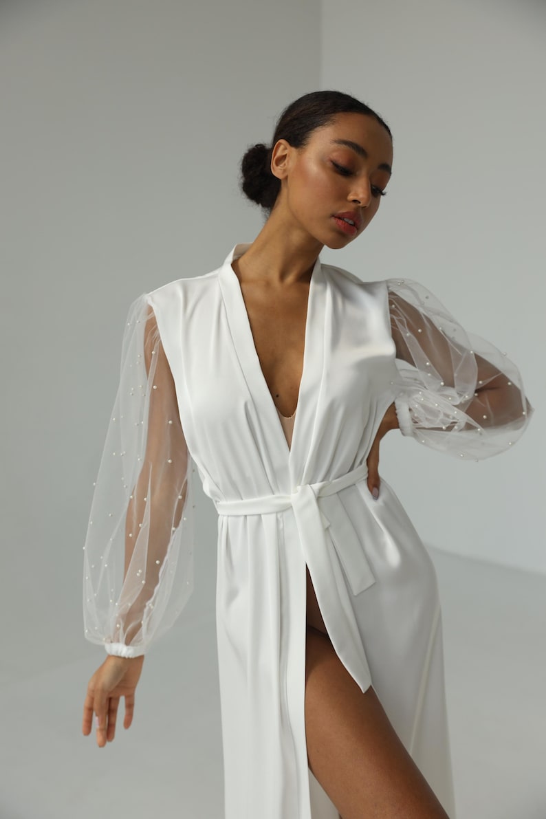 May include: A white satin robe with sheer, beaded sleeves. The robe has a deep V-neckline and a tie belt.