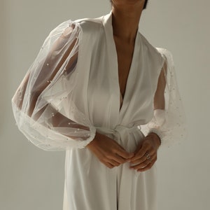May include: A white satin robe with sheer, beaded sleeves. The robe has a V-neckline and a sash tie at the waist.