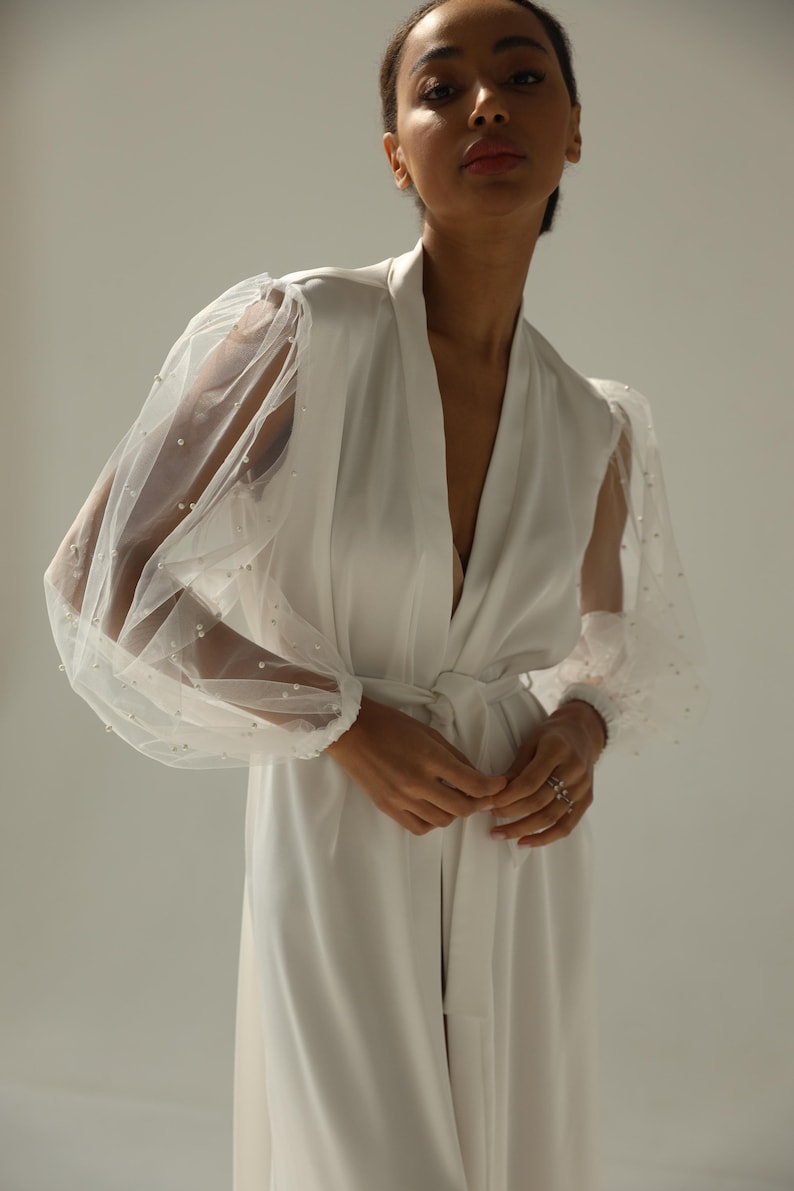 May include: A white satin robe with sheer, beaded sleeves. The robe has a V-neckline and a sash tie at the waist.