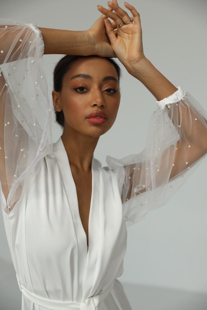 May include: A white robe with a deep V-neckline and long sleeves. The sleeves are made of sheer fabric with pearl embellishments.