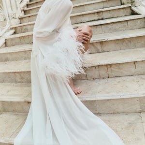 May include: A white, flowing robe with long sleeves and feather trim. The robe is worn on a stone staircase. The sleeves are adorned with white feathers. The robe's fabric drapes down the stairs.