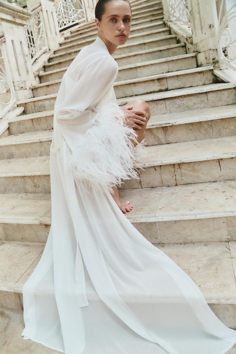 May include: A white, flowing robe with long sleeves and feather trim. The robe is worn on a stone staircase. The sleeves are adorned with white feathers. The robe's fabric drapes down the stairs.
