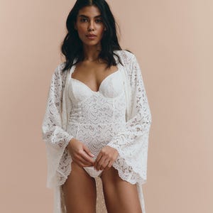 May include: A white lace robe with long sleeves and a deep V-neckline. The robe is made of delicate lace and has a flowing silhouette.