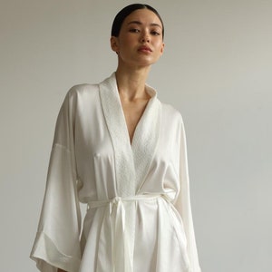 May include: A white satin robe with a white trim around the collar and sleeves. The robe has a sash that ties at the waist.