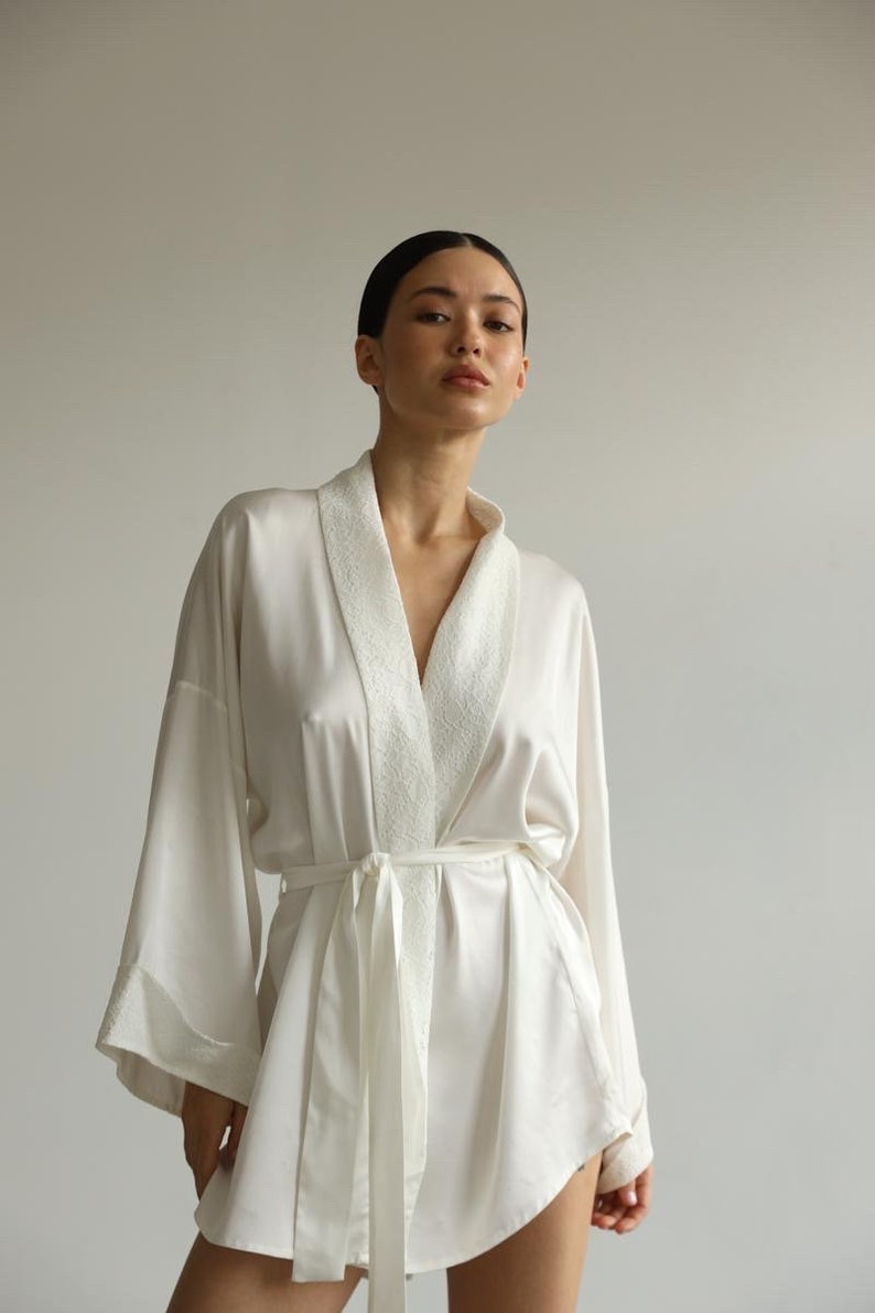 May include: A white satin robe with a white trim around the collar and sleeves. The robe has a sash that ties at the waist.