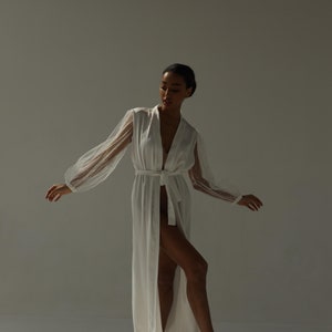 May include: A white satin robe with sheer polka dot sleeves and a sash tie. The robe is open and the model is wearing white fluffy slippers.