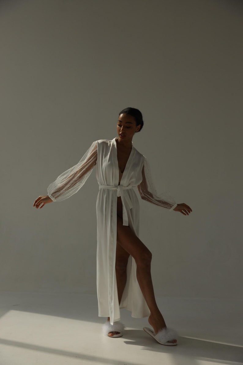 May include: A white satin robe with sheer polka dot sleeves and a sash tie. The robe is open and the model is wearing white fluffy slippers.