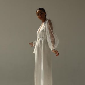 May include: A white robe with a long, flowing skirt and sheer, dotted sleeves. The robe has a sash at the waist and is worn with white fluffy slippers.