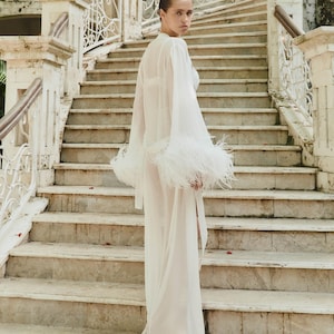 May include: A sheer white robe with long sleeves and feather trim on the cuffs. The model is standing on a marble staircase with ornate railings. The background features a stone building and palm trees, creating a luxurious aesthetic.