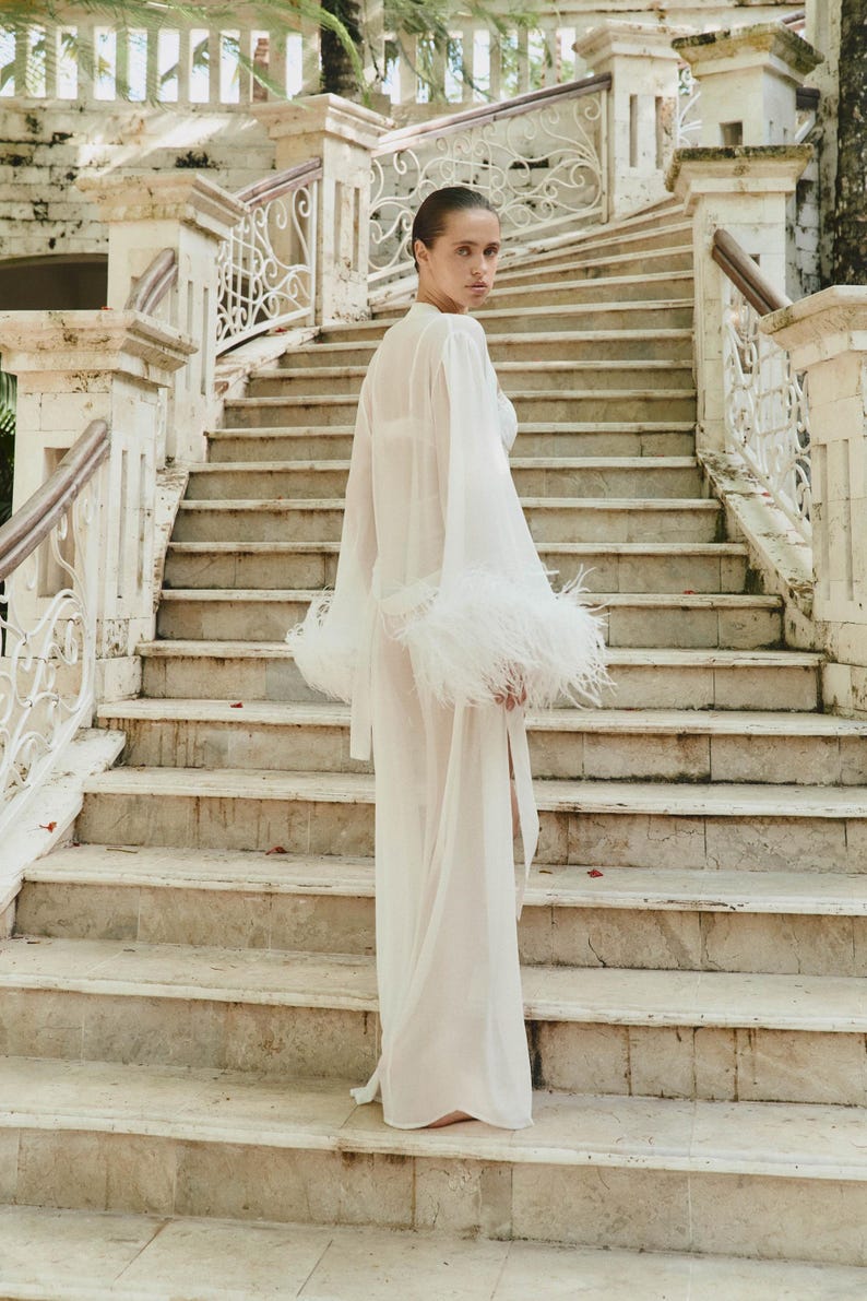 May include: A sheer white robe with long sleeves and feather trim on the cuffs. The model is standing on a marble staircase with ornate railings. The background features a stone building and palm trees, creating a luxurious aesthetic.