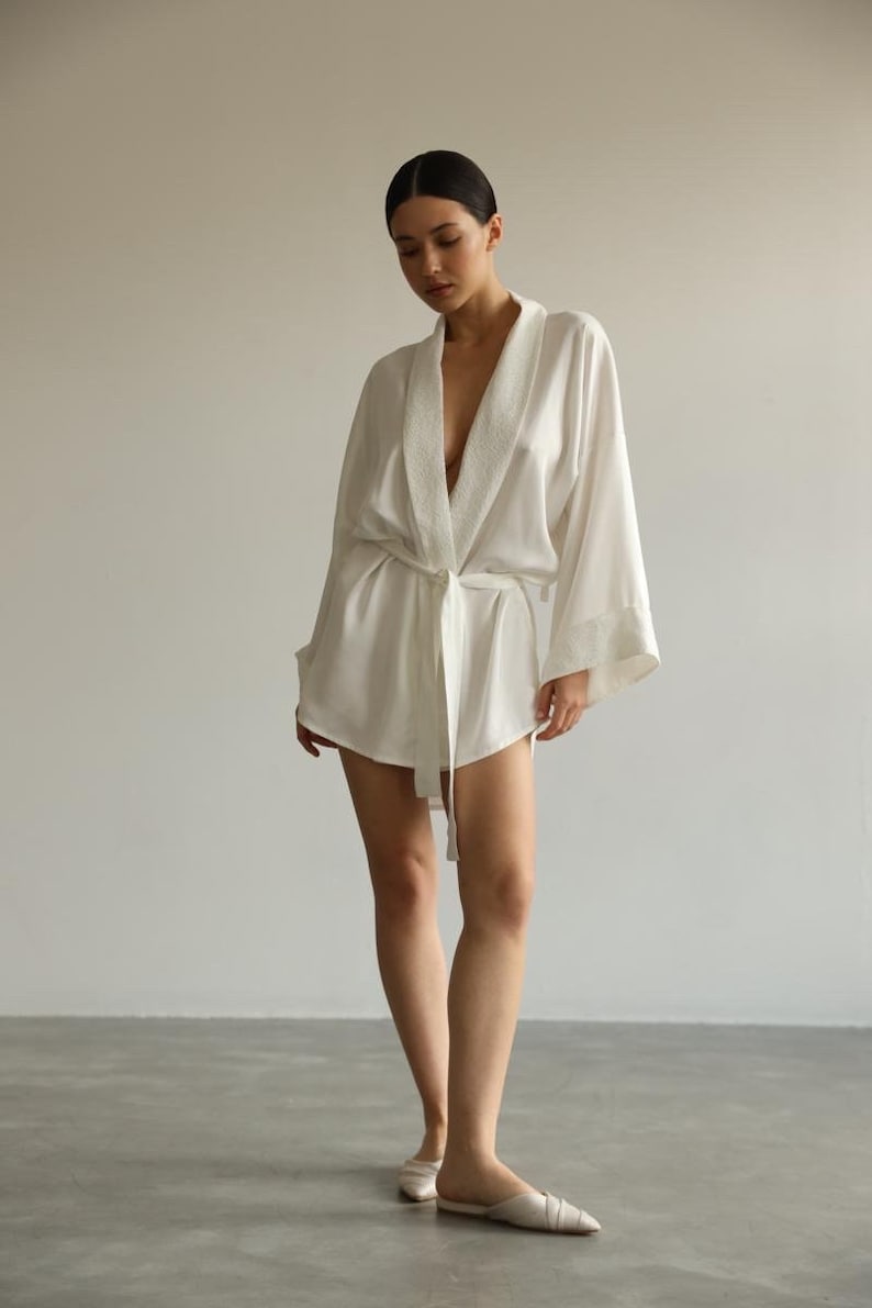 May include: A white satin robe with a shawl collar and a tie belt. The robe has a short length and wide sleeves. The robe is made of a soft, silky fabric.