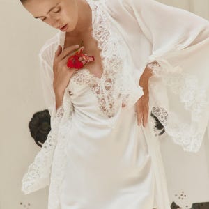 Könnte beinhalten: Weißes Satin-Dessous-Set mit Slipdress und einem transparenten Morgenmantel mit Spitzenbesatz. Eine rote Blume setzt einen Farbakzent. Das Set ist auf Komfort und Eleganz ausgelegt.